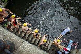 In Badekleidung steigen die Schwimmer:innen die Treppe zur Spree hinunter. Einer von ihnen macht einen Köpfer.