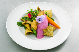 Ein kunstvoll angerichteter, vegetarischer Gourmetteller aus der taz Kantine: In der Mitte eines großen, weißen Tellers liegt ein farbenfrohes Arrangement aus gefüllten Teigtaschen, gebratenem Gemüse, jungem Blattspinat und einer ganzen kleinen Karotte. G