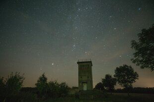 Der alte Grenzturm steht umgeben von Bäumen in der Landschaft, über ihm wölbt sich der Sternenhimmel