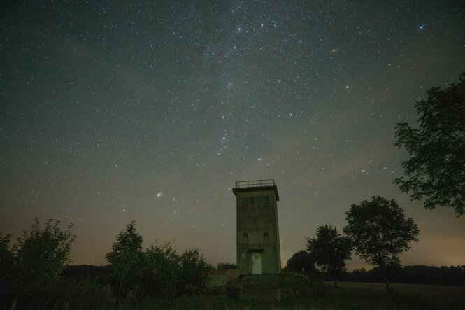 Der alte Grenzturm steht umgeben von Bäumen in der Landschaft, über ihm wölbt sich der Sternenhimmel