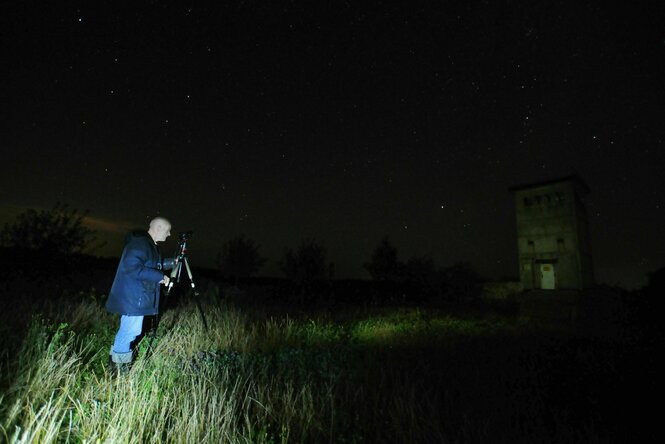 Der Fotograf steht mit seiner Kamera in der Nacht, er wird angestrahlt von einer seitlichen Lichtquelle