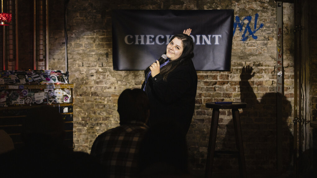 Rebecca Pop - lange dunkle Haare, dunkle Jacke, gestreifte Bluse steht auf einer Bühne, hinter ihr eine Backsteinwand an der ein Banner mit der Aufschrift "Checkpoint" hängt