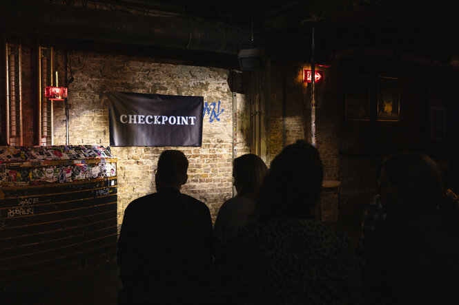 Blick vom Publikum auf die Bühne - Backsteinwand an der ein Banner mit der Aufschrift "Checkpoint" hängt