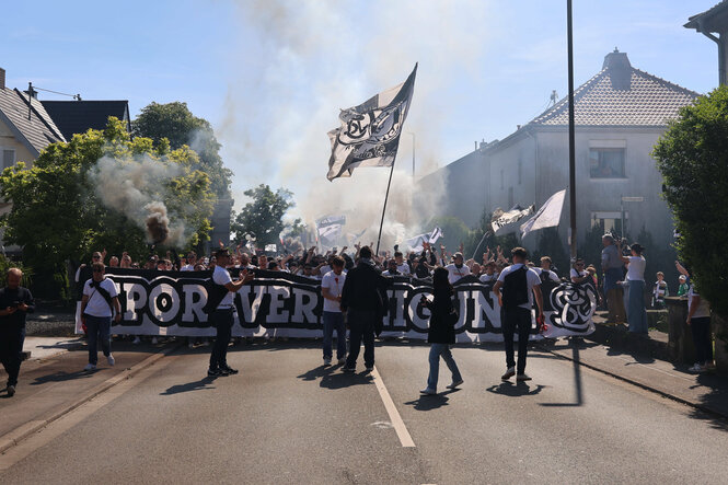 Fußballfans laufen mit einem großen Banner, Fahnen und Rauchbengalos durch einen Ort