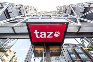 Blick von unten auf das moderne Gebäude der taz mit Stahl- und Glaselementen. Im Vordergrund hängt ein rotes Schild mit dem weißen „taz“-Logo und einer Tatzenpfote über dem Eingang. In den Glasscheiben spiegeln sich benachbarte Häuser.