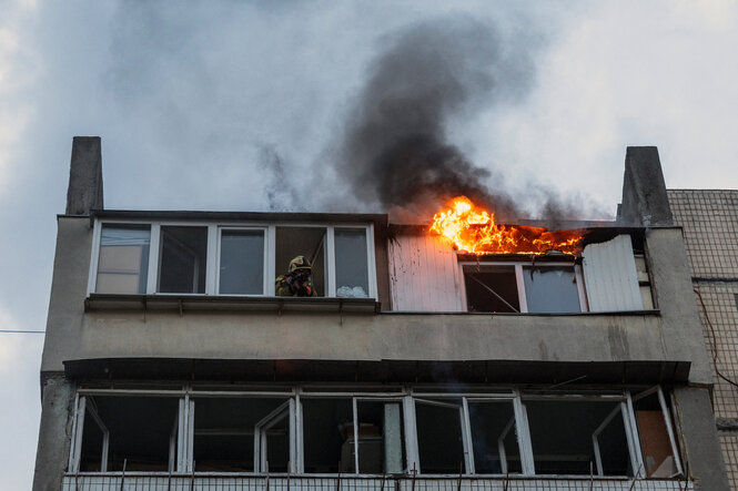Es brennt über einem Fenster in einem Wohnhaus. Aus einem anderen Fenster im selben Stockwerk schaut ein Feuerwehrmann heraus