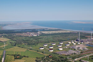 Luftaufnahme von Industrieanlagen mit mehreren großen Tanks, die sich in einer grünen Landschaft befindet. Zwischen ihnen ist ein Naturschutzgebiet. Im Hintergrund ist ein ausgedehnter Küstenbereich mit Meer und Sandbänken zu sehen