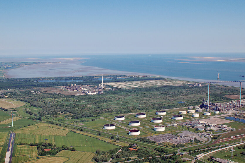 Luftaufnahme von Industrieanlagen mit mehreren großen Tanks, die sich in einer grünen Landschaft befindet. Zwischen ihnen ist ein Naturschutzgebiet. Im Hintergrund ist ein ausgedehnter Küstenbereich mit Meer und Sandbänken zu sehen