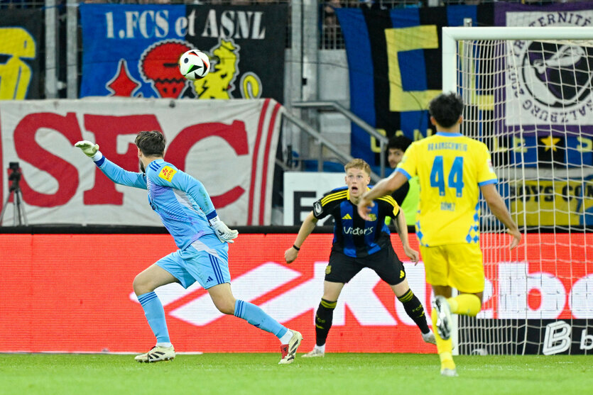 Ein Torwart in blauem Trikot versucht, einen Schuss eines angreifenden Spielers in Gelb vor dem Tor zu blocken, während ein Verteidiger daneben steht.