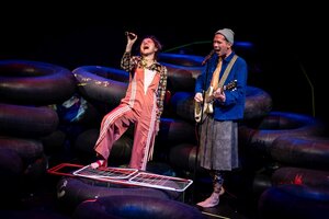 Ein Mann im Kleid mit Mütze an der Gitarre, die Frau singend mit Mikrofon im Arbeitsanzug stehen vor Autoreifen