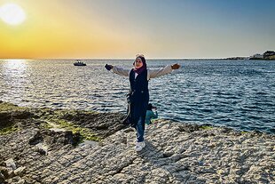 Mehr als ein Gefühl von Freiheit: Eine Frau mit lila Hijab und Sonnenbrille steht mit ausgebreiteten Armen auf einer felsigen Küste und blickt in die Kamera. Die Sonne geht links über dem glitzernden Meer unter. Ein Boot ist auf dem Wasser, und eine weitere Person ist hinter ihr auf den Felsen zu sehen.