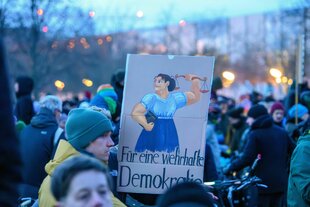 Jugendliche auf einer Demonstration mit einem Plakat: Der Slogan lautet: Für eine wehrhafte Demokratie und zeigt eine junge Frau, die ihre Muskeln spielen lässt
