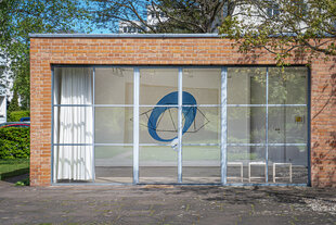Eine Außenansicht des Mies van der Rohe Haus. Durch das große Fenster sieht man eine Arbeit von May Bill mit einem blauen Kreis.