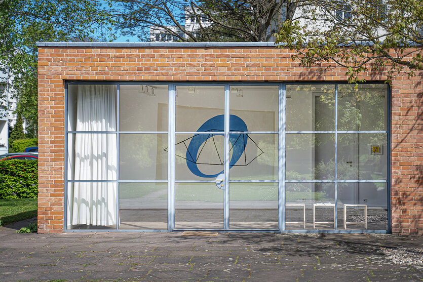 Eine Außenansicht des Mies van der Rohe Haus. Durch das große Fenster sieht man eine Arbeit von May Bill mit einem blauen Kreis.