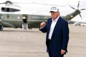 Donald Trump steht auf einem Abflugfeld vor einem Helicopter, trägt eine MAGA Basecap und ballt die Faust