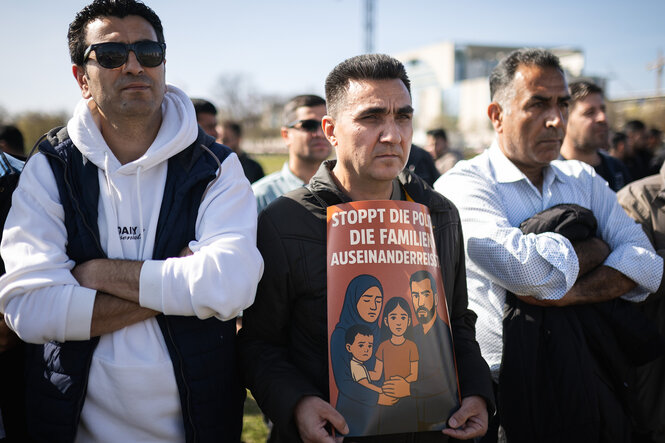 Männer protestieren stumm, einer hält ein Plakat mit der Illustration einer Familie