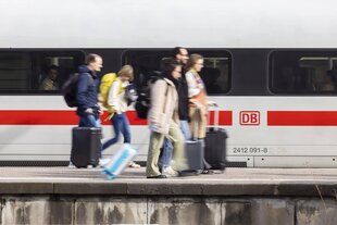 Menschen mit KOffern gehen auf einem Bahnsteig an einem ICE entlang. Sie sind in Bewegungsunschärfe abgebildet