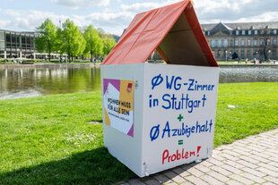 Schöner Wohnen? – Ein kleines Modellhaus aus Pappe mit orangefarbenem Dach steht auf einer Wiese vor einem See. Darauf steht: „Ø WG-Zimmer in Stuttgart + Ø Azubigehalt = Problem!“.