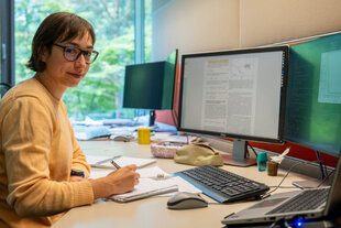 Hin- und hergerissen zwischen Heidelberg und Lissabon: Beatriz Bordadágua, eine junge Frau mit Brille und hellorangem Pullover, sitzt an ihrem aufgeräumten Schreibtisch und macht sich Notizen. Vor ihr stehen zwei Monitore mit wissenschaftlich anmutenden Inhalten. Durch ein Fenster im Hintergrund blickt man ins Grüne.
