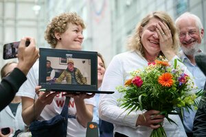 Eine Person hält einen Blumenstrauß und lacht, eine andere Hält neben ihr einen Laptop hoch, auf dem jemand zugeschaltet ist