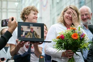 Eine Person hält einen Blumenstrauß und lacht, eine andere Hält neben ihr einen Laptop hoch, auf dem jemand zugeschaltet ist