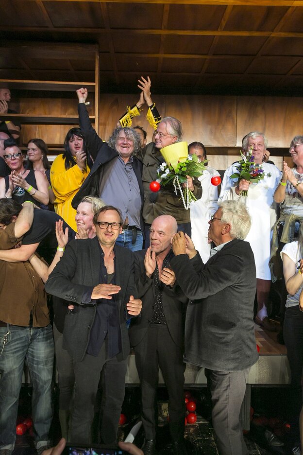 Carl Hegemann steht beim Schlussapplaus uaf de Bühne zwischenScauspielern und SChauspielerinnen und reisst die Arme hoch