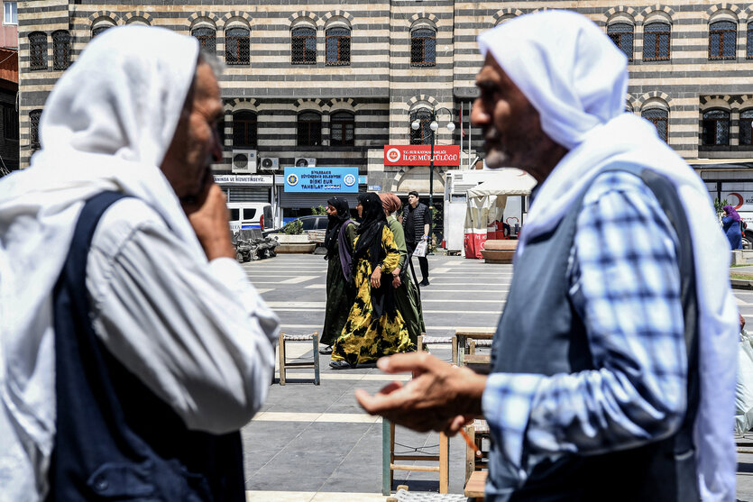 Zwei Personen stehen auf einem Platz und unterhalten sich, im Hintergrund läuft eine Gruppe Frauen entlang, alle tragen Tücher um den Kopf