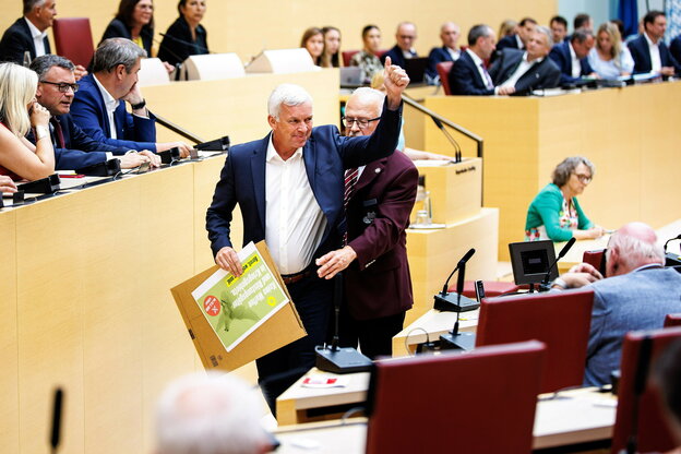 Ralf Stadler wird von einer Person durch die Reihen eines Plenarsaals geführt, er streckt den Daumen seiner Hand nach oben