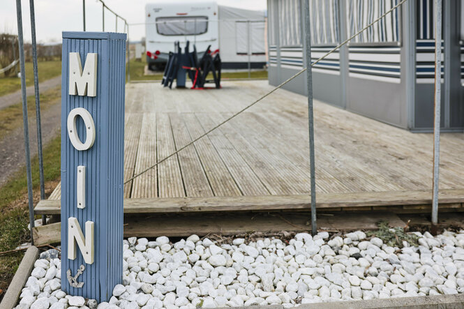 Ein Pfeiler mit der Aufschrift "Moin" steht vor einem Wohnwagen auf dem Campingplatz Ostseecamping in Schubystrand.