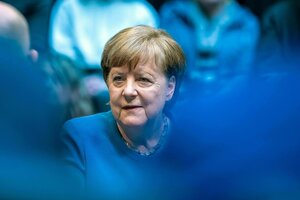 Angela Merkel steht zwischen blau gekleideten Menschen. Auch die ist blau gekleidet. Weil vor ihr, also näher an der Kamera, zwei Menschen in Blau stehen oder sitzen, von denen man nur die Arme und Schultern sieht,, sieht Merkel sehr klein aus.