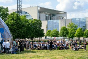 Die knapp 40 Teilnehmenden des "Parlaments der Menschen" sitzen in einer Reihe und hören einem Redner zu, im Hintergrund das Kanzleramt