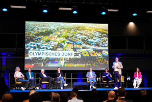 Podium mit Menschen vor einer Projektion "Olympisches Dorf"
