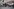 Trauernde stehen vor einem Haus mit Fotos von Kindern und Erwachsenen, im Hintergrund Brandspuren