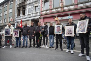 Trauernde stehen vor einem Haus mit Fotos von Kindern und Erwachsenen, im Hintergrund Brandspuren