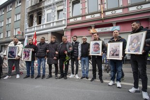 Trauernde stehen vor einem Haus mit Fotos von Kindern und Erwachsenen, im Hintergrund Brandspuren