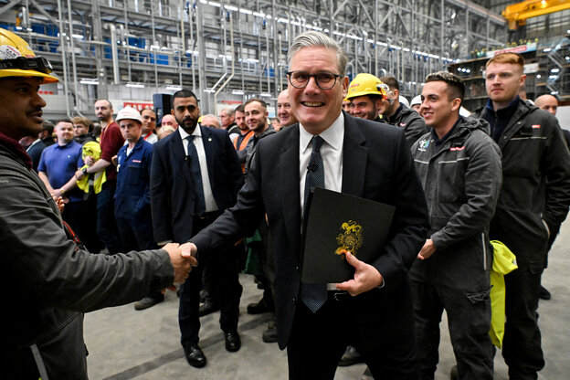 Keir Starmer wendet sich von einem Arbeiter mit Schutzhelm ab zur Kamera, sie stehen zwischen einer Menge in einer Fabrikhalle