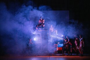 Eine Gruppe Männer mit nackten Oberkörpern versammelt um Musikboxen und Bierkästen