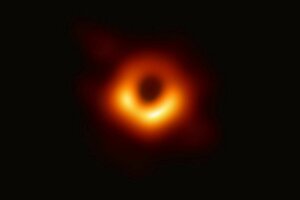 Das Bild zeigt das erste Foto eines Schwarzen Lochs, ein dunkler, runder Bereich in der Mitte, umgeben von einem hellen, orangefarbenen Lichtring auf schwarzem Hintergrund, das durch die Gravitation des Lochs verzerrt wird.