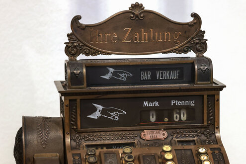 Das Bild zeigt eine antike Registrierkasse aus Metall mit der Aufschrift „Ihre Zahlung“ und „Bar Verkäufe“ in Weiß. Sie hat ein Display mit „0,60“ in Mark und Pfennig sowie runde Tasten mit Ziffern und Ornamenten.