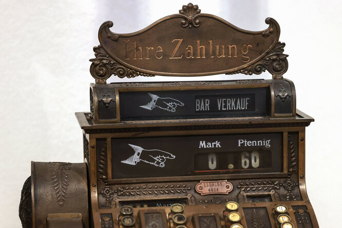 Das Bild zeigt eine antike Registrierkasse aus Metall mit der Aufschrift „Ihre Zahlung“ und „Bar Verkäufe“ in Weiß. Sie hat ein Display mit „0,60“ in Mark und Pfennig sowie runde Tasten mit Ziffern und Ornamenten.