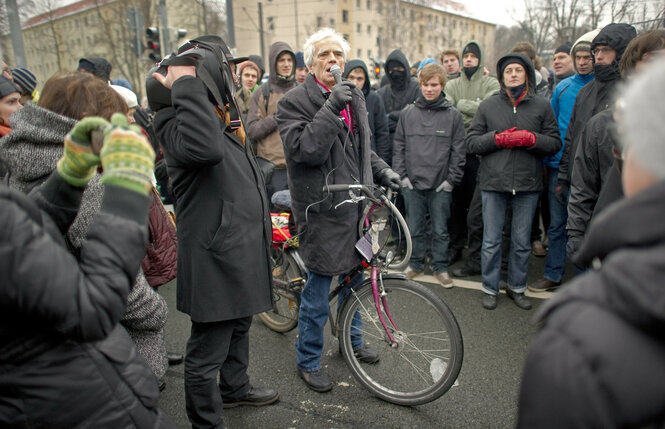 Christian Ströbele spricht bei einer Demonstration in ein Mikrofon. Er steht mit seinem Kettler-Fahrrad inmitten einer Gruppe von Demonstrierenden in Winterkleidung. Die Stimmung wirkt ernst.