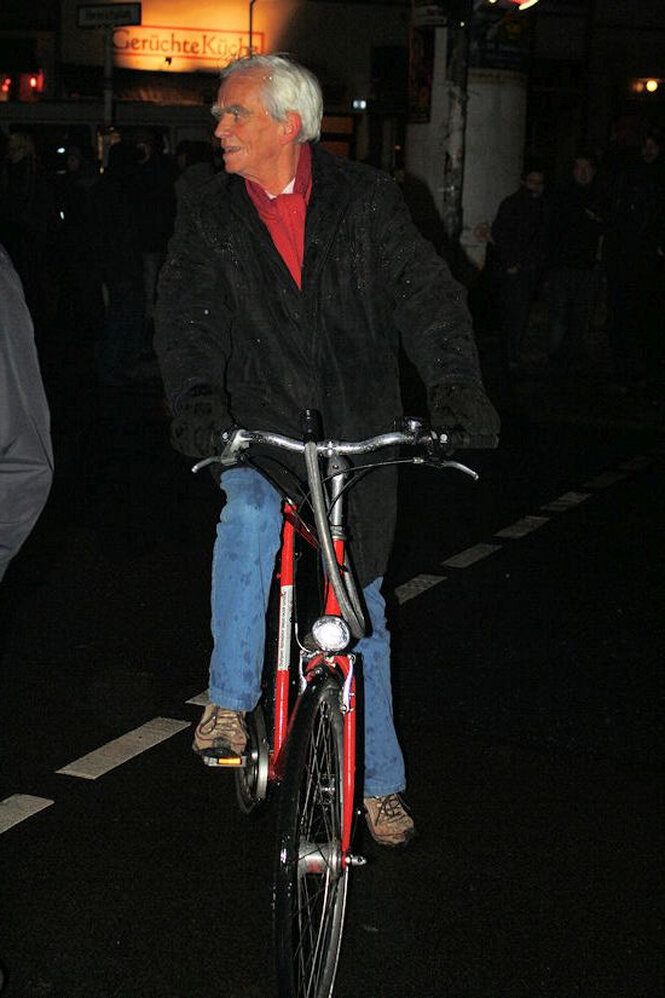 Christian Ströbele fährt bei Nacht mit einem roten Fahrrad über eine nasse Straße. Er trägt eine dunkle Jacke, Jeans und blickt zur Seite. Im Hintergrund sind verschwommen einige Menschen und ein Schild mit der Aufschrift „Gerüchteküche“ zu sehen.