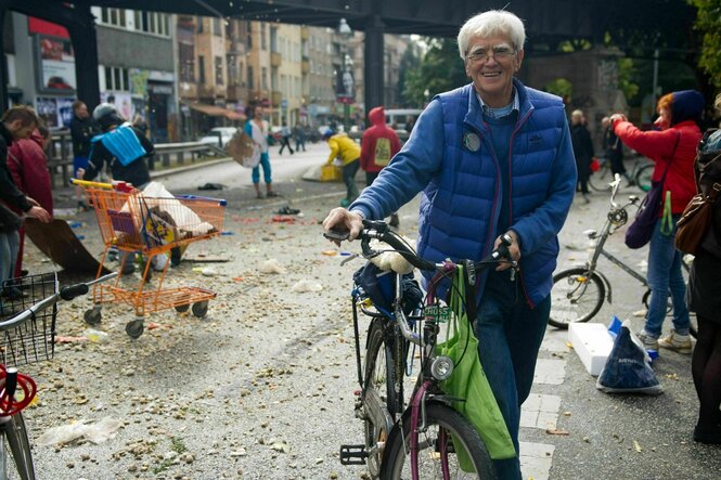 Christian Ströbele steht lächelnd mit seinem Fahrrad auf einer von Müll, Gemüseresten und Einkaufswagen übersäten Straße in Kreuzberg. Er trägt eine blaue Steppweste, Jeans und eine Brille. Im Hintergrund räumen Menschen auf, fotografieren oder unterhalten sich.
