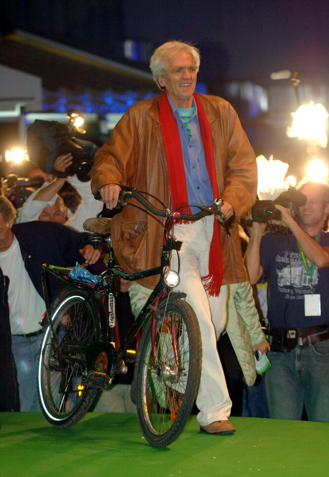 Christian Ströbele, weißhaarig, lächelt in die Menge, während er sein Kettler-Fahrrad über einen grünen Teppich schiebt. Er trägt einen braunen Leder-Mantel, einen rote Schal und helle Hosen. Um ihn herum fotografieren und filmen zahlreiche Menschen. Es ist Abend, im Hintergrund brennen Scheinwerfer. Es ist eine Szene wie auf einer Filmgala mit Ströbele als Star.