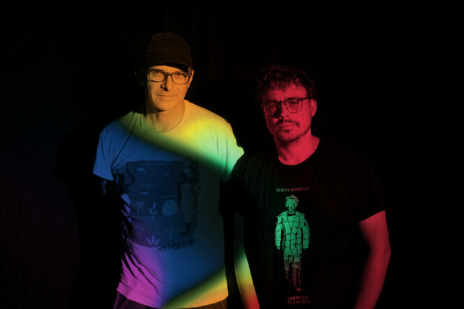 Die Musiker Florian Zimmer und Andi Haberl stehen im Dunkeln. Auf sie fällt buntes Licht in Regenbogenfarben. Beide tragen eine Brille.