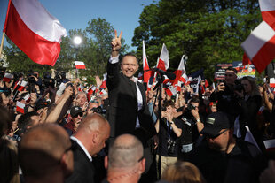 Karol Nawrocki steht in einer Menschenmenge und zwischen rot-weißen Fahnen, er macht das Victory-Zeichen