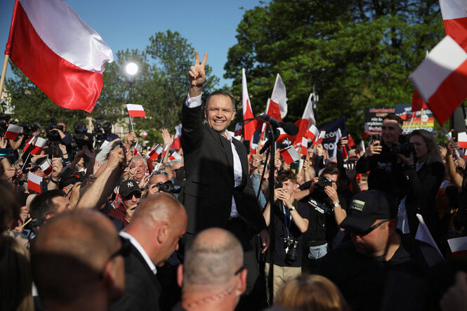 Karol Nawrocki steht in einer Menschenmenge und zwischen rot-weißen Fahnen, er macht das Victory-Zeichen