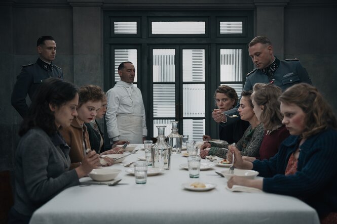 Eine Vorkosterin an der Tafel wird von einem Mann in Uniform gefüttert