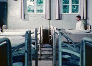 Ein kahles Zimmer mit symmetrisch angeordneten Betten. Auf einem davon sitzt eine Person.