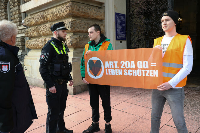 Zwei Männer halten ein Transparent mit der Aufschrift "Art. 20 A GG = Leben schützen" hoch, vor ihnen steht ein Polizist in Uniform